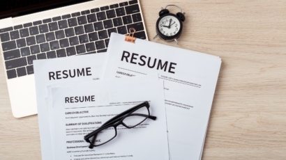 Two resumes stacked on desk with laptop, clock and glasses