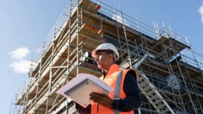 Man holding sheet of construction bids on clipboard, looking over job site with building and blue sky in the background