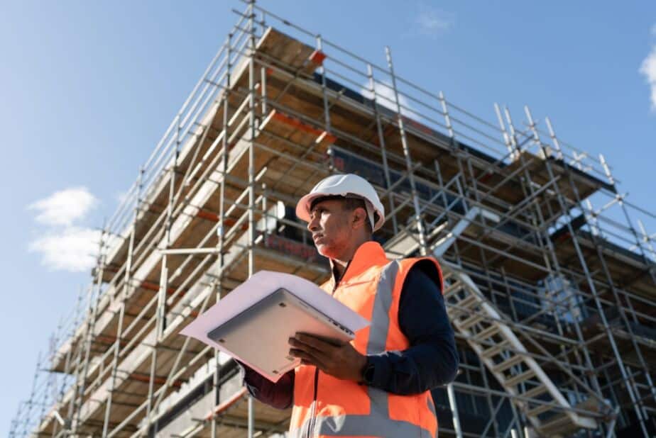 Man holding sheet of construction bids on clipboard, looking over job site with building and blue sky in the background